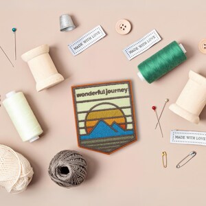 May include: A brown felt patch with a sunset and mountain scene. The patch has the text "wonderful journey" in white lettering. The patch is surrounded by sewing supplies, including spools of thread, buttons, pins, and a thimble.