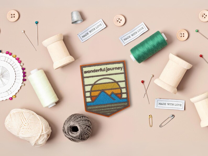 May include: A brown felt patch with a sunset and mountain scene. The patch has the text "wonderful journey" in white lettering. The patch is surrounded by sewing supplies, including spools of thread, buttons, pins, and a thimble.