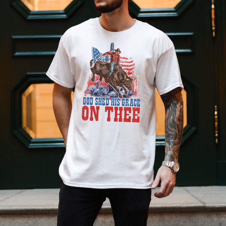 May include: White t-shirt with a graphic of a cowboy riding a bull in front of an American flag. The text "God Shed His Grace On Thee" is printed below the graphic.