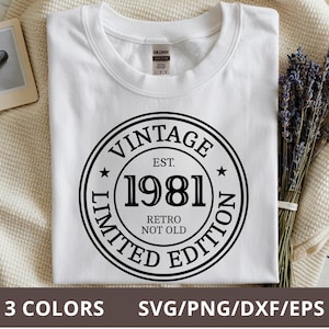 Pode incluir: Camiseta branca com um gráfico circular preto que diz "VINTAGE EST. 1981 RETRO NOT OLD LIMITED EDITION". A camisa está dobrada sobre um cobertor creme, com lavanda e uma pequena foto emoldurada.