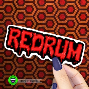 以下が含まれることがあります： 赤い塗料が黒の背景に垂れている「Redrum」のビニールステッカー。ステッカーは赤とオレンジのパターンのある表面に貼られています。