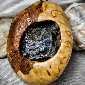 Puede incluir: Colgante artesanal con una piedra oscura y texturizada engastada en un marco de madera. La madera tiene un acabado natural de color claro por un lado y un acabado más oscuro y teñido por el otro. Cordón de cuero marrón.