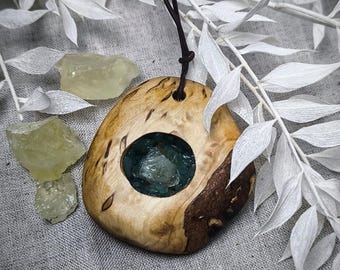 May include: A wooden pendant with a circular cutout filled with green and blue crystals. The pendant is attached to a brown leather cord.