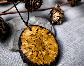 May include: Oval wooden pendant with a natural wood grain pattern and a dark brown border. The pendant hangs from a brown leather cord. The background includes pine cones and a gray stone.
