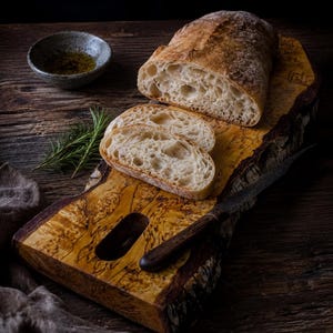 May include: A loaf of crusty bread, partially sliced, rests on a wooden cutting board with a live edge. Two slices of bread are in front. A small bowl of olive oil and a sprig of rosemary are also present. A dark knife is on the board.