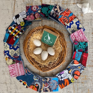 May include: A circle of 14 colorful zippered pouches with various African print patterns. The pouches are arranged around a woven basket with eggs inside. A small wooden box with the text "Adventure Trade Shop" is in the center of the basket.