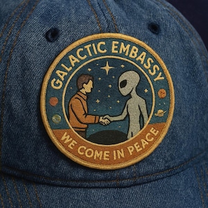 Könnte beinhalten: Dunkelblaue Jeans-Baseballkappe mit einem runden Aufnäher. Der Aufnäher zeigt die Aufschrift "GALACTIC EMBASSY" und "WE COME IN PEACE" und zeigt einen Menschen und einen Außerirdischen, die sich die Hände schütteln. Der Hintergrund ist ein Sternenhimmel.