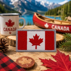 May include: A rectangular patch featuring the Canadian flag with a red maple leaf on a white background, framed by red bars. A white mug with a red maple leaf and the word "CANADA" is also visible. A red canoe and other Canadian themed items are in the background.