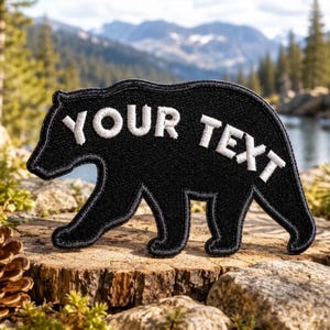 May include: A black bear-shaped embroidered patch featuring the white stitched text "YOUR TEXT" across the center. The patch has a thick black border and a textured fabric surface, designed to be applied to clothing or other textile items.