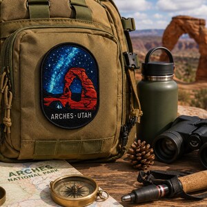 Op de afbeelding: Een beige tactische rugzak met een ovaal geborduurde patch die de rode rotsboog van Arches National Park onder een sterrenhemel weergeeft, met de tekst ARCHES UTAH in het wit onderaan het ontwerp van de patch.