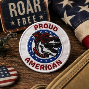 May include: A round, white patch with the words "PROUD AMERICAN" in red embroidery. The patch features a dinosaur design with red and black details, surrounded by blue stars. Additional patriotic items are in the background.