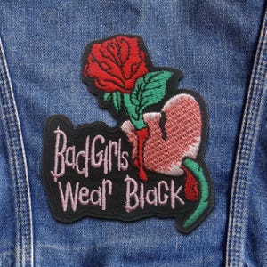 May include: Embroidered patch with a red rose, a broken pink heart, and the text "Bad Girls Wear Black" in pink. The patch has a black border and green details. It is attached to a blue denim jacket.