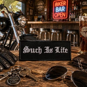 May include: A black patch with the embroidered text "Such Is Life" sits on a wooden table, alongside a motorcycle, sunglasses, keys, a wallet, a glass of cola, and a pair of black leather gloves. A neon sign reads "Biker Bar Open".