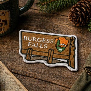 May include: A Burgess Falls patch with a brown wooden sign design and white border. The patch features the text "BURGESS FALLS" and a park service logo. The patch is on a wooden surface with a compass, mug, and jacket.
