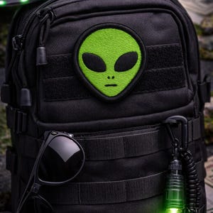 May include: Black tactical backpack with a green alien patch. The backpack features multiple compartments, a pair of sunglasses, and a glowing green flashlight. A miniature UFO is visible in the background, suggesting a space or sci-fi theme.
