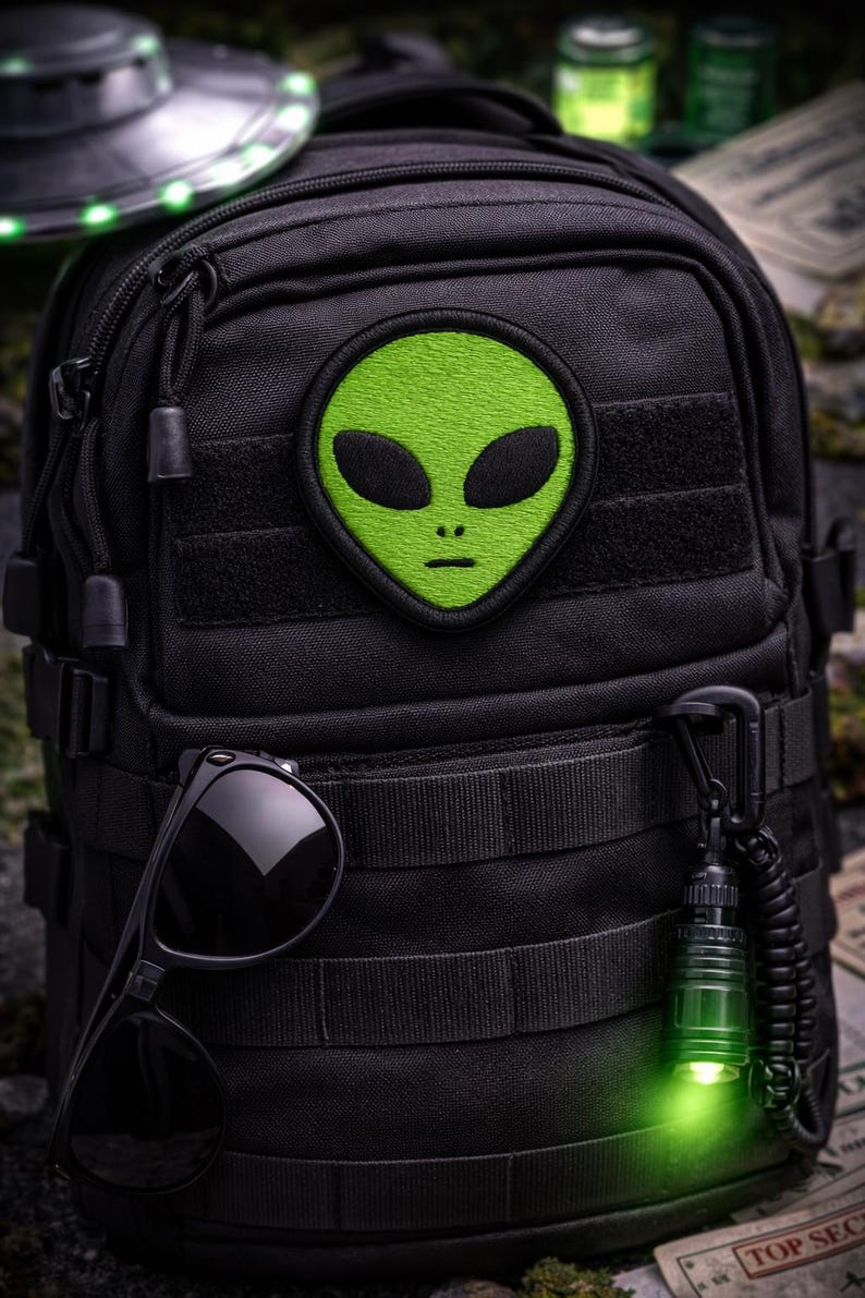 May include: Black tactical backpack with a green alien patch. The backpack features multiple compartments, a pair of sunglasses, and a glowing green flashlight. A miniature UFO is visible in the background, suggesting a space or sci-fi theme.