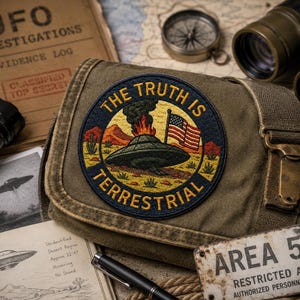 Op de afbeelding: Een ronde geborduurde patch met een donkergroene UFO die neerstort in een woestijnlandschap met een Amerikaanse vlag en rook, omringd door de tekst THE TRUTH IS TERRESTRIAL, bevestigd aan een bruine canvas schoudertas met metalen gesp.