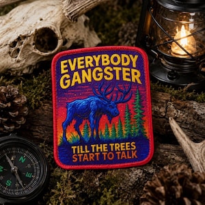 May include: A rectangular embroidered patch with a red border featuring a blue moose standing in a forest against a sunset background. The text reads EVERYBODY GANGSTER TILL THE TREES START TO TALK in yellow and orange lettering on a purple field.