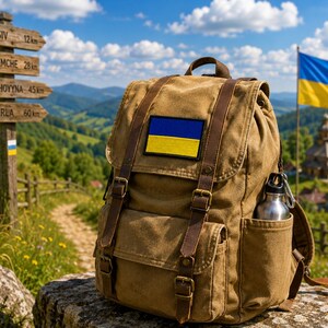 May include: A brown canvas backpack with leather straps and a Ukrainian flag patch. A stainless steel water bottle is in the side pocket. The background includes a wooden signpost, a Ukrainian flag, and a scenic mountain view.