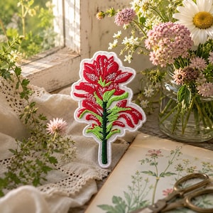 May include: Embroidered patch featuring a red and green flower design with a white border. The patch is placed on a cream-colored surface near a window with a bouquet of flowers and a vintage book.