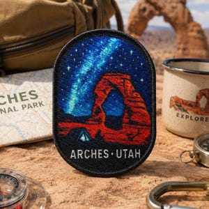Peut inclure: Un écusson brodé ovale représentant un ciel nocturne avec une nébuleuse bleue brillante et des étoiles au-dessus d'une arche rocheuse rouge. L'écusson a une bordure noire et le texte blanc ARCHES UTAH imprimé en bas, illustrant un paysage désertique.