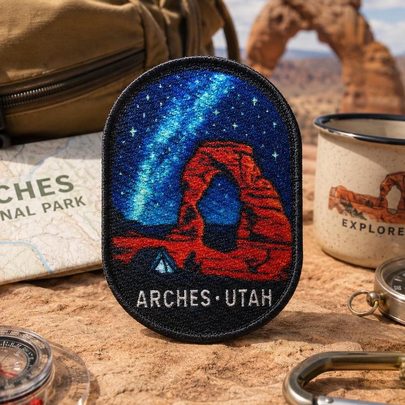 Op de afbeelding: Een ovaal geborduurde patch met een nachtelijke hemel met een helderblauwe nevel en sterren boven een rode rotsboogformatie. De patch heeft een zwarte rand en de witte tekst ARCHES UTAH onderaan, wat een woestijnlandschap weergeeft.