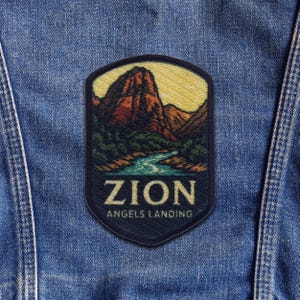 以下が含まれることがあります： ザイオン国立公園の風景を描いたパッチ。川、山、そして「ZION ANGELS LANDING」の文字があります。パッチは濃い青色の縁取りがあり、デニムの背景に配置されています。