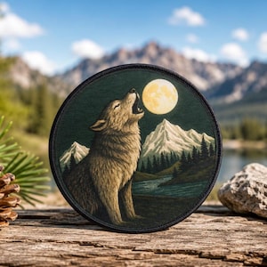 Könnte beinhalten: Ein runder bestickter Aufnäher mit einem braunen Wolf, der den vollen gelben Mond anheult. Die Szene zeigt schneebedeckte Berge, einen dunklen Nadelwald und einen Fluss, der durch die Landschaft fließt, alles umrandet von einem schwarzen Saum.