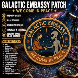 Puede incluir: Un parche bordado circular con un fondo espacial azul oscuro con estrellas y planetas. El diseño muestra a una persona con camisa naranja estrechando la mano de un alienígena gris. El texto dice GALACTIC EMBASSY y WE COME IN PEACE.