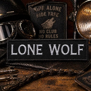 May include: A black embroidered patch with the words "LONE WOLF" in white block letters. The patch is set against a backdrop of motorcycle gear, including a helmet, goggles, and a headlight.