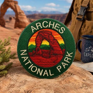 May include: A circular embroidered patch featuring a dark green border with the white text ARCHES NATIONAL PARK. The center depicts a red rock arch formation against a sunset sky with yellow, orange, and red horizontal bands above green hills.