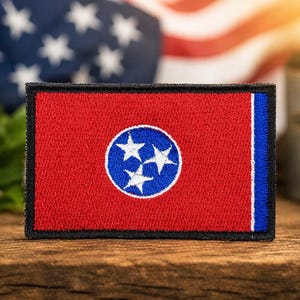 May include: A rectangular patch featuring the Tennessee state flag, with a red background, a blue vertical stripe, and a central blue circle containing three white stars. The patch is on a wooden surface with an American flag in the background.