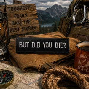 May include: An outdoor adventure scene with a lit lantern, compass, map, and a patch that reads "BUT DID YOU DIE?". A wooden sign says "TOUGHEST TRAILS MAKE THE BEST STORIES". A mug with "SURVIVED ANOTHER ADVENTURE" sits next to a backpack and rope.
