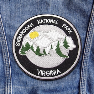 May include: A circular embroidered patch featuring a black border with the white text SHENANDOAH NATIONAL PARK and VIRGINIA. The center depicts a white bear silhouette filled with green pine trees and a yellow sun against a light gray background.