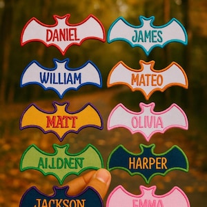 Op de afbeelding: Geborduurde vleermuisvormige patches met namen als Daniel, James en William. Elke patch heeft een ander kleurenschema, waaronder rood, blauw en geel, met de namen in contrasterende kleuren. De patches worden in een hand gehouden tegen een vage achtergrond.