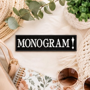 May include: A black rectangular patch with the word "MONOGRAM!" embroidered in white. The patch is surrounded by a beige handbag, a floral notebook, sunglasses, and a woven hat.