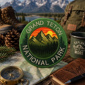 May include: A circular embroidered patch featuring a green border with the text GRAND TETON NATIONAL PARK. The center depicts a mountain range in shades of green under a bright yellow sun and orange sky, with a dark green forest in the foreground.