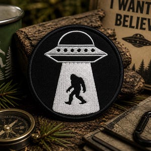 May include: A black and white embroidered patch featuring a UFO beaming up a Bigfoot silhouette. The patch is round and has a black border. Other items in the image include a compass, a mug with a Bigfoot design, and a sign that says "I WANT TO BELIEVE."