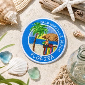 May include: A round, blue embroidered patch with the text "Jacksonville Beach Florida". The patch features a palm tree, beach scene, and a bar with a thatched umbrella. The patch is surrounded by seashells, driftwood, and sand.