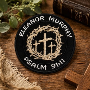 May include: A black circular patch with white text reading "ELEANOR MURPHY" and "PSALM 91:11". The patch features a gold-colored cross design within a gold-colored thorny wreath. The patch is on a wooden surface.