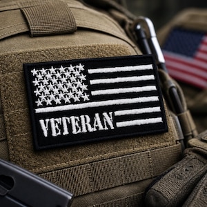 May include: A black and white embroidered patch with the word "VETERAN" and a stylized American flag design. The patch is affixed to a tan tactical vest, with a partial view of a firearm and the American flag in the background.