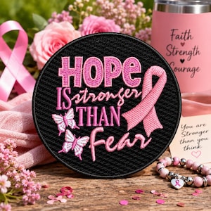 May include: A circular black embroidered patch featuring the pink text HOPE IS stronger THAN fear, accompanied by two white and pink butterflies and a pink awareness ribbon. The design is stitched with a textured finish on a dark fabric base.