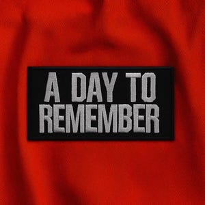 Könnte beinhalten: Ein schwarzer, rechteckiger Aufnäher mit dem weißen Schriftzug "A DAY TO REMEMBER". Der Aufnäher ist vor einem leuchtend roten Stoffhintergrund platziert.