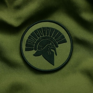 Puede incluir: Un parche circular verde oliva con un diseño de casco espartano verde oscuro. El parche está bordado con una ilustración detallada del casco, sobre un fondo verde oliva liso. El parche está cosido sobre una tela verde oliva.