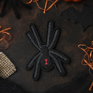 May include: A black spider patch with gray stitching and a red hourglass marking. The patch is surrounded by Halloween decorations, including an orange pumpkin, a black bat, and orange ribbons. The background is a dark, textured surface.