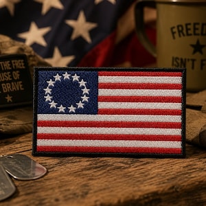 May include: Embroidered patch featuring the Betsy Ross flag design, with a blue canton of 13 white stars in a circle and red and white stripes. The patch is rectangular with a black border. The background includes military-themed items.