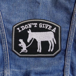 May include: A black patch with white embroidered text and imagery. The text reads "I DON'T GIVE A". The image depicts a donkey being led by a rat. The patch is sewn onto a blue denim jacket.
