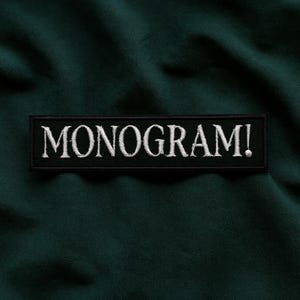 Pu&ograve; includere: Sfondo in tessuto verde scuro con una toppa rettangolare nera. La toppa presenta la scritta "MONOGRAM!" ricamata in bianco, con un carattere serif. Il tessuto ha un aspetto leggermente strutturato.