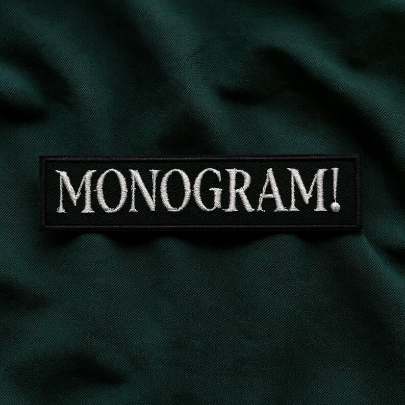 Pu&ograve; includere: Sfondo in tessuto verde scuro con una toppa rettangolare nera. La toppa presenta la scritta "MONOGRAM!" ricamata in bianco, con un carattere serif. Il tessuto ha un aspetto leggermente strutturato.
