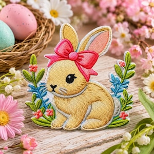 May include: An embroidered patch featuring a light brown bunny with a bright pink bow on its head, surrounded by green leaves, small red flowers, and blue floral accents, designed with a detailed stitched texture and a clean white border.